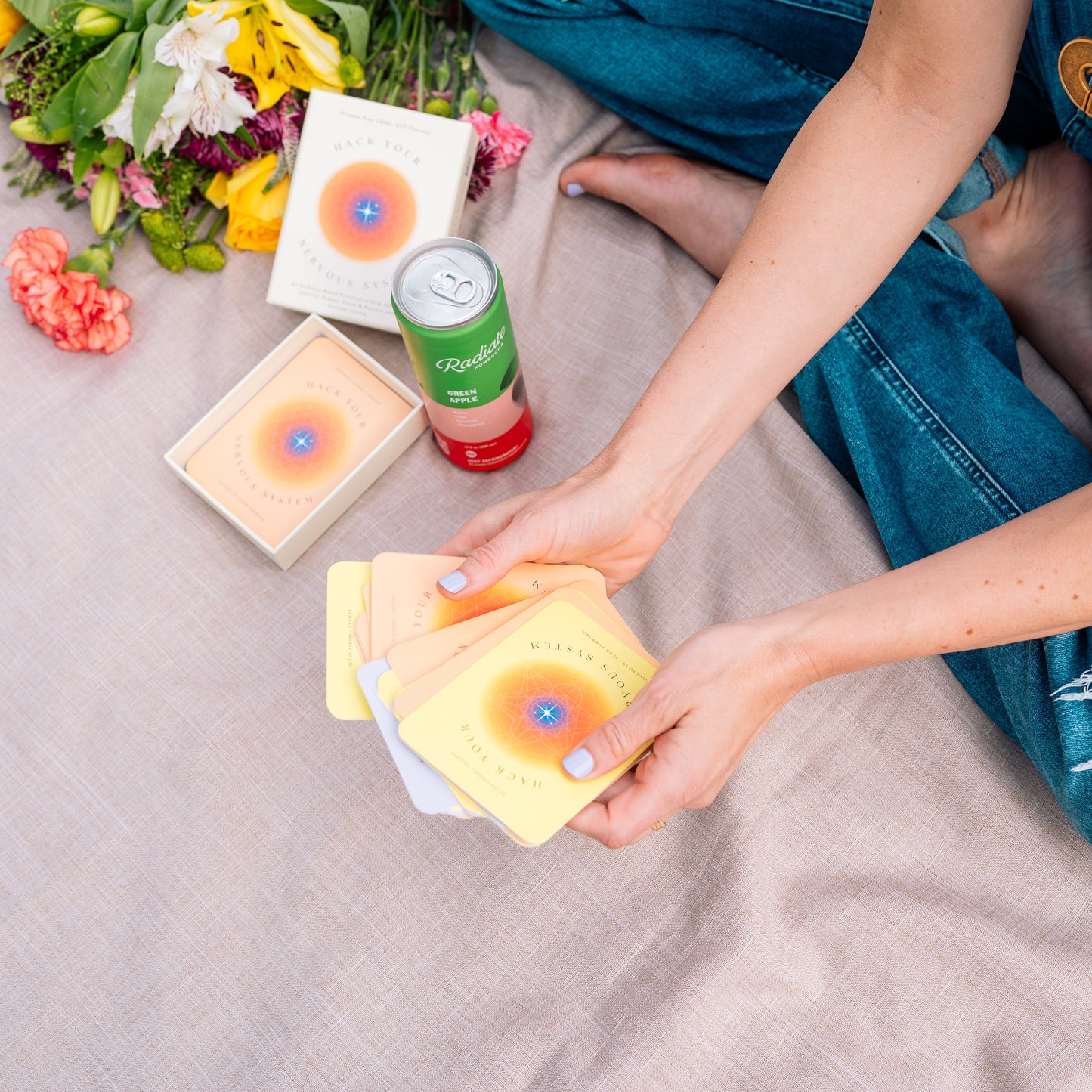 Person sitting on a blanket with colorful cards and a drink, surrounded by flowers.